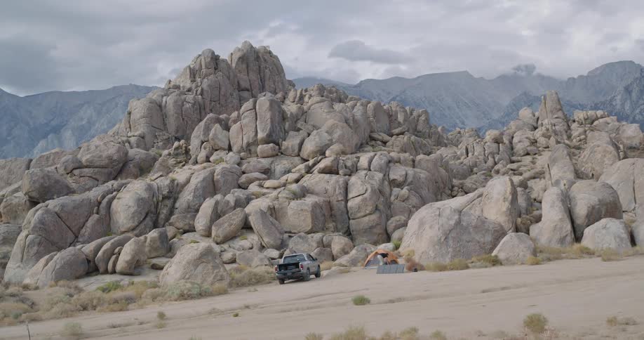 A man stands near a silver pickup and orange tent at a rocky desert campsite beneath towering boulders appearing calm and relaxed
