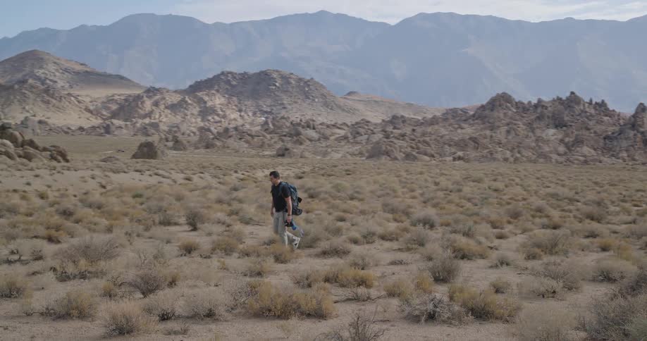Man walking through desert terrain with sparse vegetation and mountains in the background