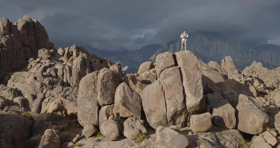 A person with a backpack moves across and then stands at the highest point of a large prominent rock formation in the foreground turning to look at the vast distant mountain ranges beneath a dark sky