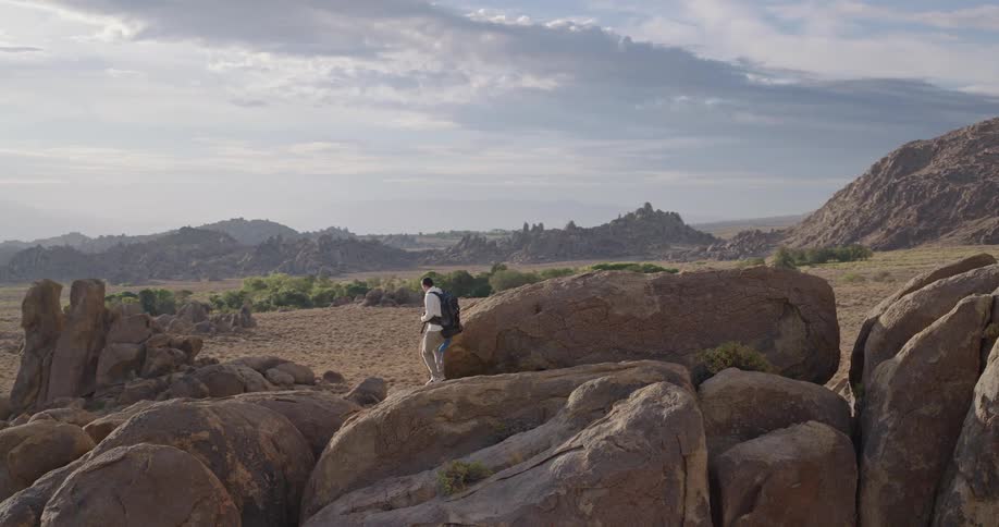 The video is an establishing shot of a vast highaltitude desert or scrubland strewn with large rounded boulders and dominated by imposing hazy mountain ranges in the backgrounda solitary hiker with a backpack stands on top of a prominen