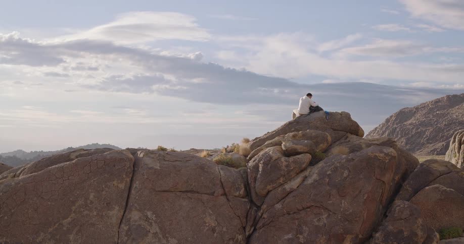 The video is an extreme long shot that slowly pans out showing a hiker sitting alone on a large prominent redbrown rock formation in a desert landscapethe wide vista is hazy with bright sunlight and features many rounded boulders and distant