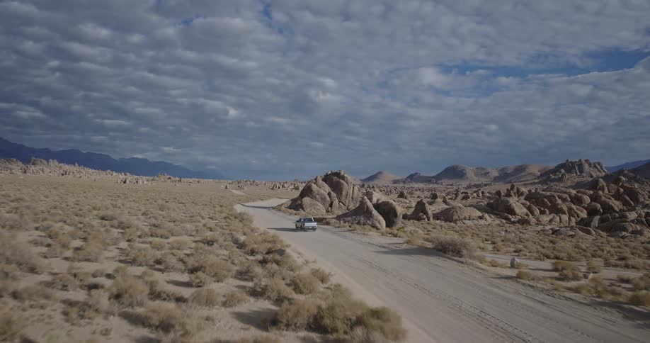 A gray suv drives away from the camera on a straight unpaved road through a wide sparsely vegetated desert