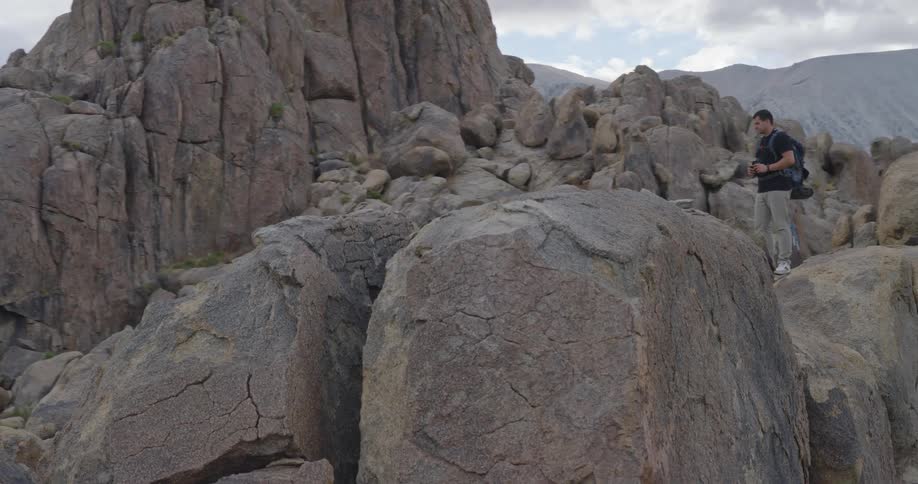 A hiker with a prosthetic leg and a backpack navigates a rocky mountainous terrain stopping to use binoculars