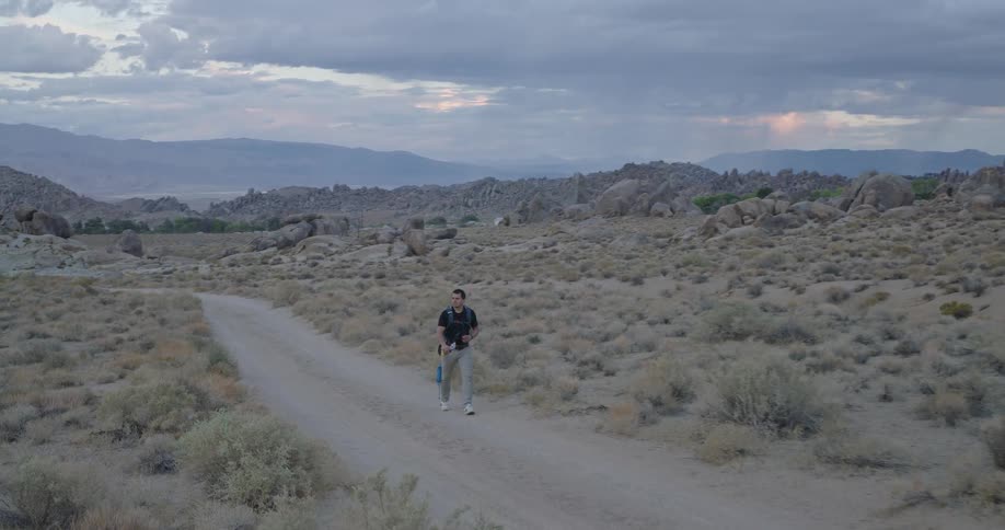 A man with a backpack hikes along a dirt road through a vast rocky desert landscape under a cloudy overcast sky