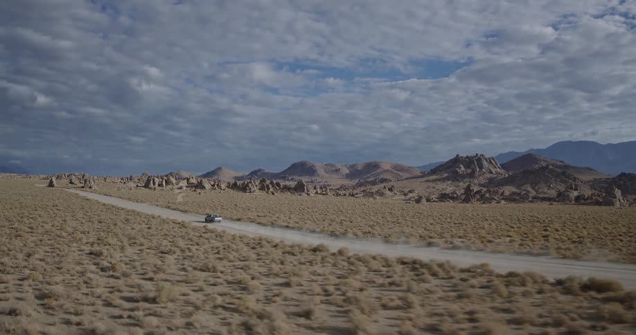 An aerial view follows a lone pickup truck driving along a dirt road through a vast rocky desert landscape under a dramatic cloudy sky