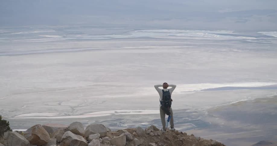 Person standing on rock with backpack overlooking salt flat surrounded by mountains