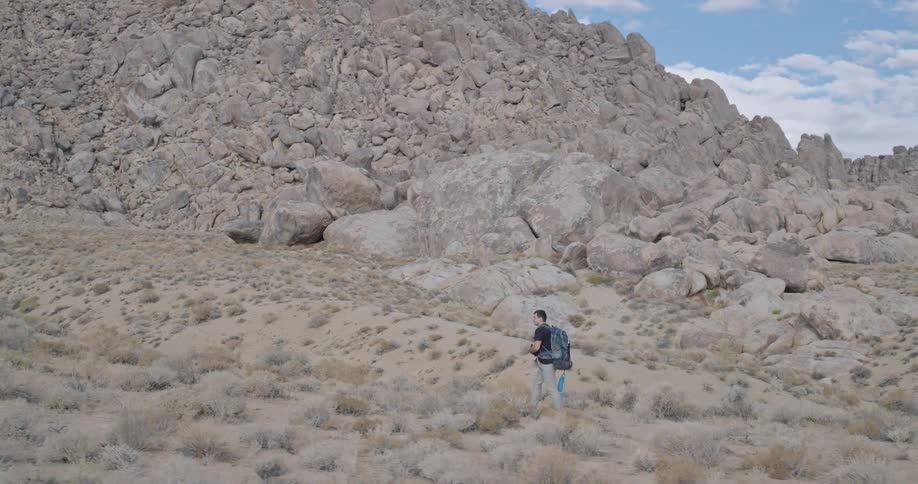 A lone hiker with a blue backpack walks across a scrubby desert valley beneath towering granite ridges appearing calm and purposeful