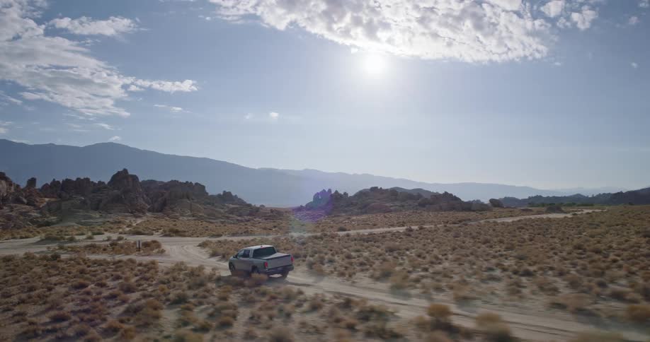 An aerial shot follows a silver pickup truck as it drives along a dusty desert road surrounded by rocky terrain and with mountains visible in the distance under a partly cloudy sky