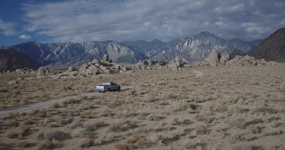 A silver pickup travels along a dusty track across a wide sagebrush plain with dramatic granite mountains under a patchy cloud filled sky