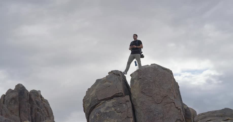 Man standing up on top of a tall rock looking around through binoculars at a mountainous region outdoors during day time