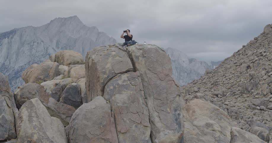 A man sits on the peak of a large prominent rock formation using binoculars to look toward a massive bright gray mountain in the far background under an overcast sky
