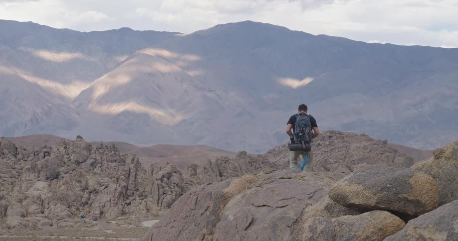 Man with backpack on rocky overlook observing mountain range under partly cloudy sky