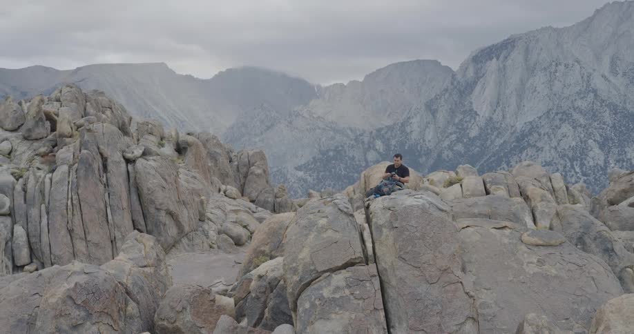 A man sits alone on a large boulder surrounded by a dramatic rocky mountain landscape under a cloudy sky