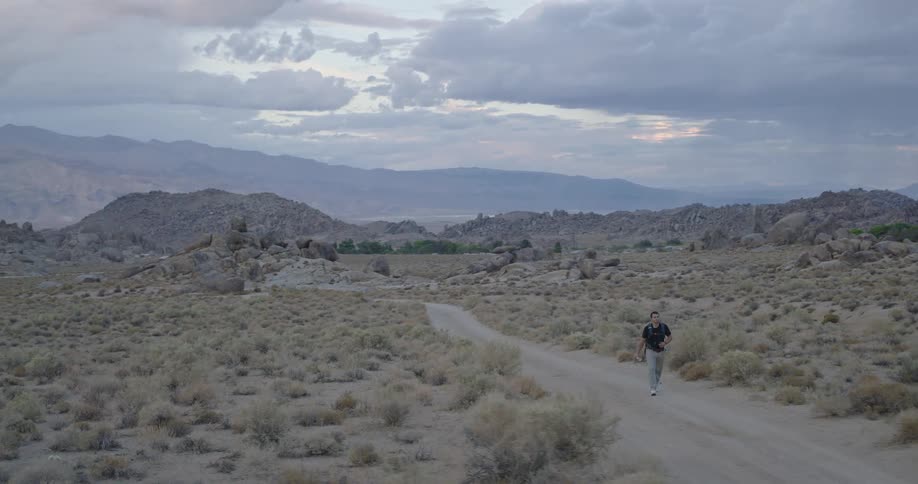 A lone man walks along a sandy desert road at dusk with layered mountains under a cloudy sky