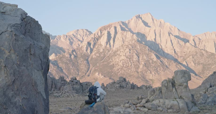 A man in a hoodie carrying a backpack sits on a cliff high up a rocky mountain in the wilderness during a sunny day hike