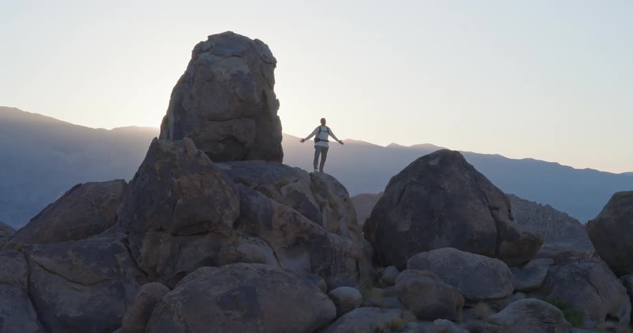 A man stands atop a prominent cylindrical rock formation with arms outstretched set within a rocky desert landscape against a backdrop of distant hills