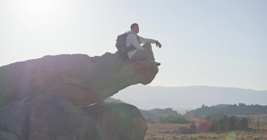 The video shows a male hiker sitting down on a large overhanging rock formation in a desert landscape he is wearing a backpack and gazing out over the expansive hazy valley and distant mountain range under bright daylight