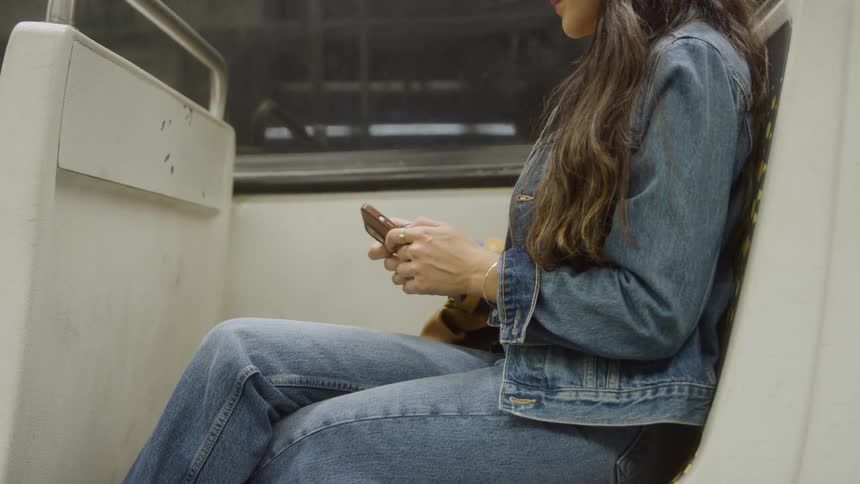 A woman wearing a denim jacket and jeans is seated inside a public transit car with white and gray walls holding a cellphone in both hands and interacting with its screen