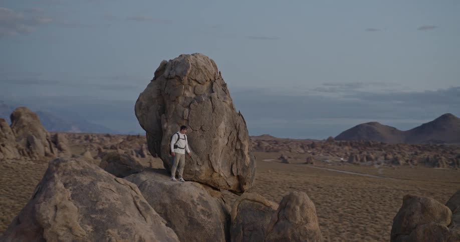 The video is an extreme long shot showing a hiker standing atop a very tall prominent rock formation in a vast arid mountainous landscapethe scene is set around sunset or sunrise with soft light casting long shadows and illuminating the