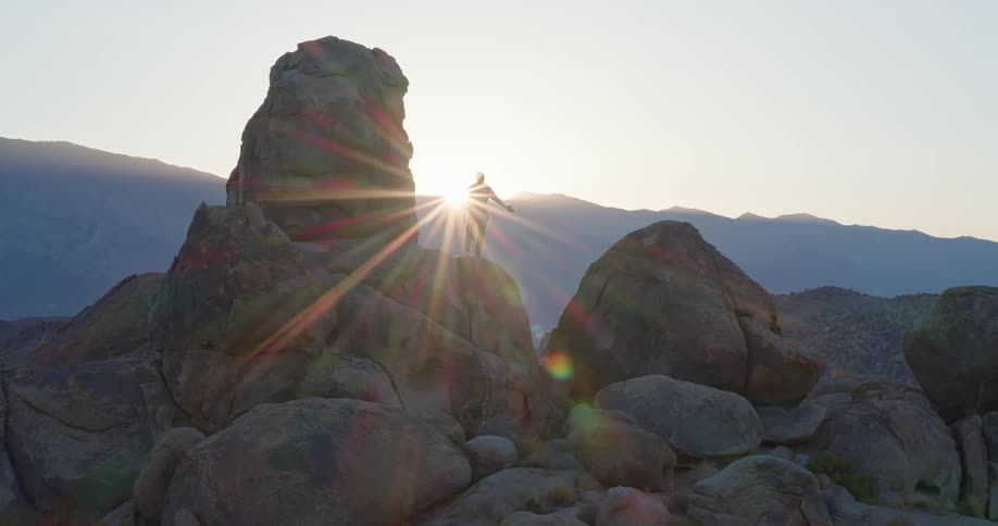 Man standing on rock formation with arms open facing mountainous landscape