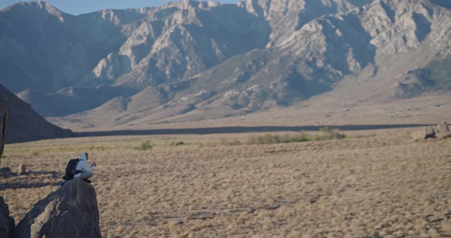 Person with backpack and hoodie sitting on rock in arid mountainous landscape interacting with device under blue sky