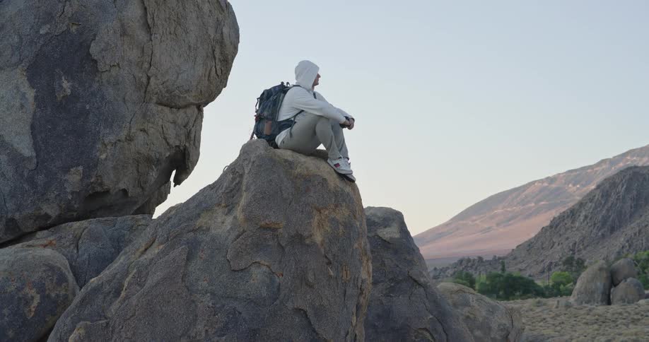 Man with a backpack sitting on a tall rock looking around at a mountainous area outdoors during dusk or dawn