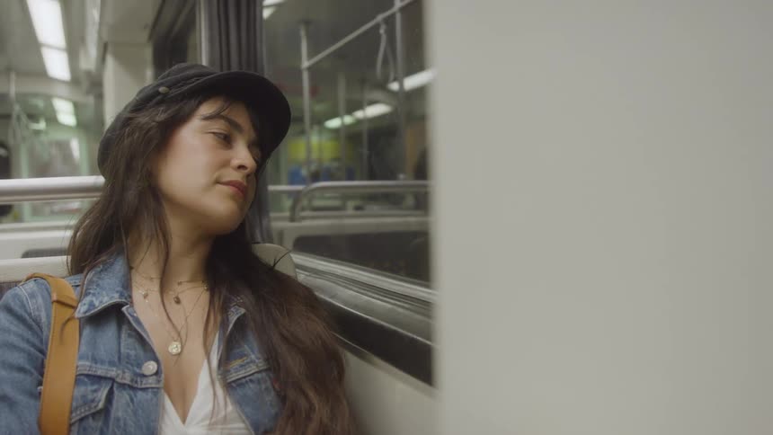 Young female wearing a black hat and a denim jacket sitting on a subway looking out of the window indoors