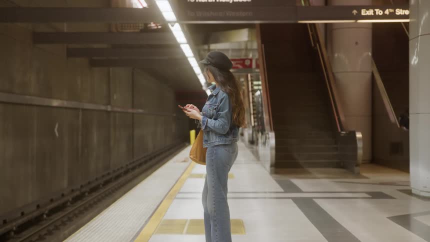 Woman standing on subway platform looking at phone with yellow bag on shoulder