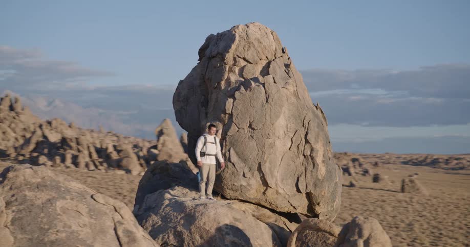 A young man hikes and climbs up rugged mountain terrain standing on a cliff and staring at the surroundings on a sunny day