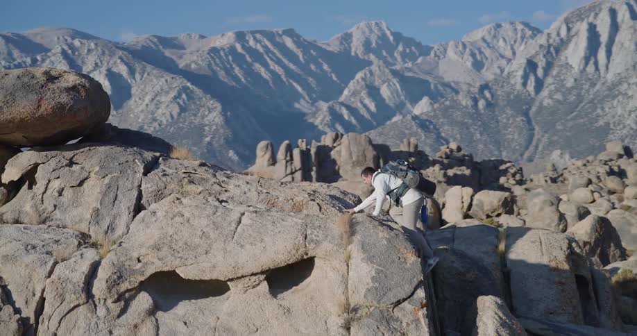 A young man hikes and climbs up a mountain on a sunny day carrying a water bottle and a backpack