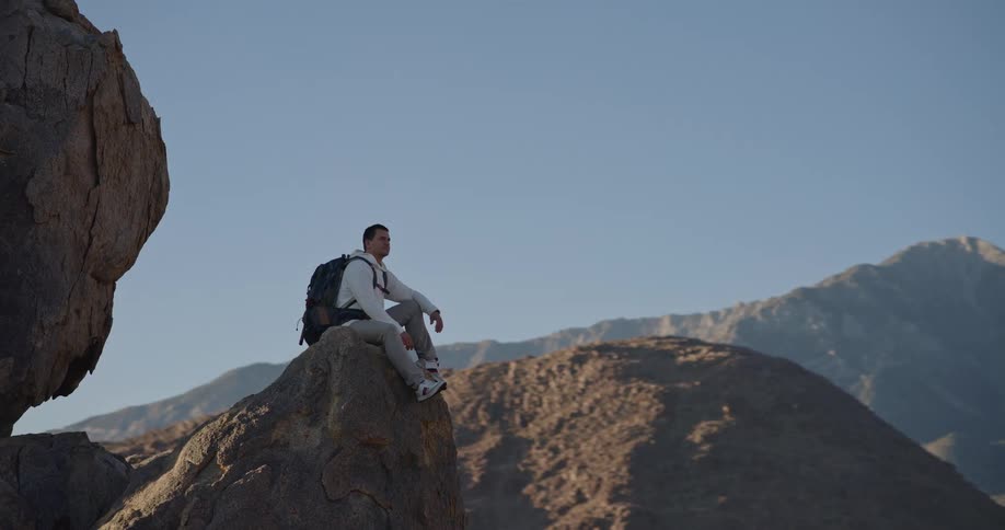 The video shows a male hiker sitting on a large jagged rock formation he is looking out at a stunning panoramic view of a massive snowcapped mountain range under a bright clear sky