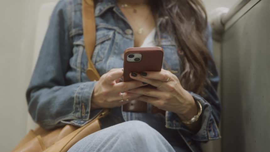 Woman with long brown hair seated in public transit wearing denim jacket and gold accessories focused on smartphone with brown case