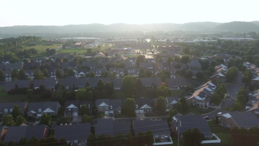 The video is an aerial long shot of a suburban residential area surrounded by rolling treecovered hills the sunlight creates a bright haze over the landscape highlighting the uniformity of the houses and the surrounding greenery