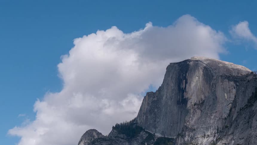 Half dome rises behind sky with white clouds above rocky cliff
