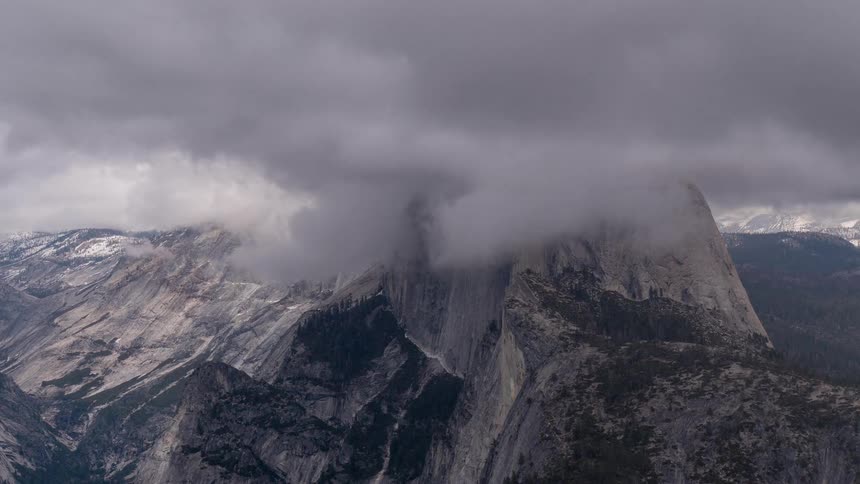 Granite cliff shrouded in low clouds rises among forested slopes