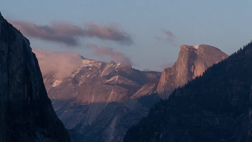 Granite cliff lit by sunlight stands out among snowy mountains and dark forest