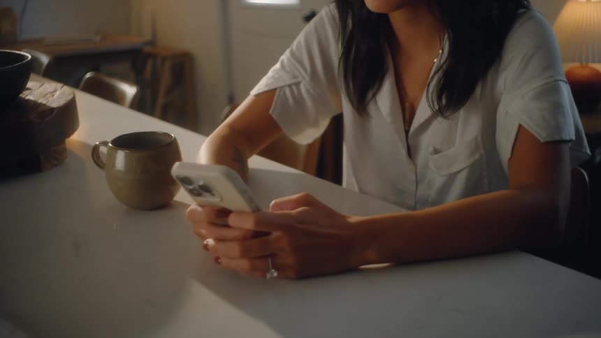 An asian woman is seated at a white counter in a dimly lit room looking down at a white mobile phone she holds in both hands with a tan coffee mug resting near her elbow on the counter