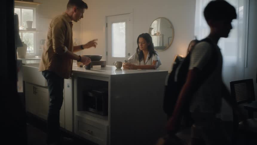 A man in a brown jacket and his two children walk down a dark staircase and enter a bright sunlit kitchen where a woman sits at a white island counter having coffee as the family prepares to start their day