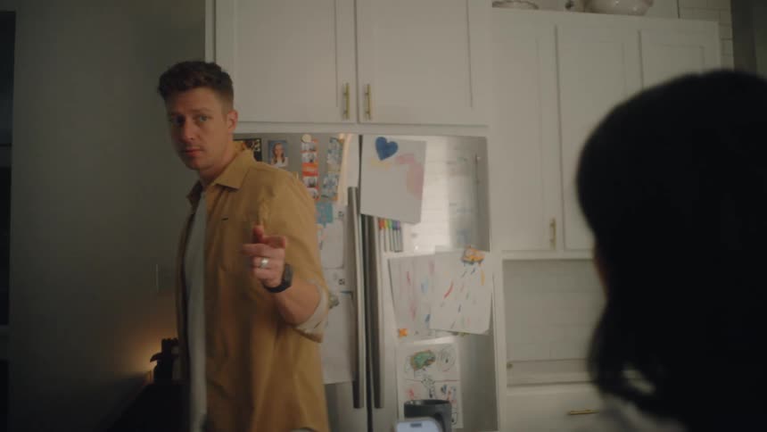 A young couple converse in the kitchen while their kids prepare to leave for school in the morning