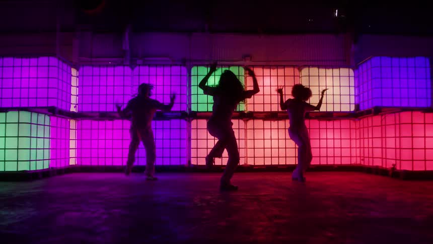 A trio of women dance with a lot of energy in front of illuminated cubes