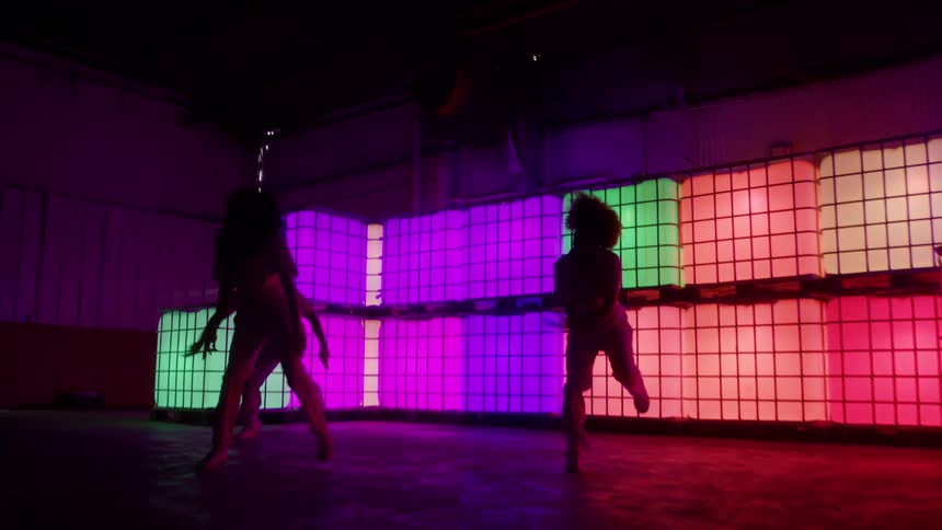 The silhouettes of three dancers perform a synchronized routine in a dark space illuminated by a large wall of glowing multicolored cubes
