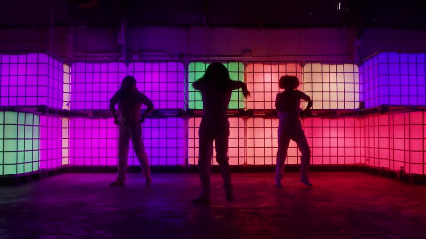 Three dancers are silhouetted against a wall of glowing brightly colored square panels that shift between red orange green and purple as they perform a choreographed routine with dynamic arm movements in a dark studio or nightclub setting