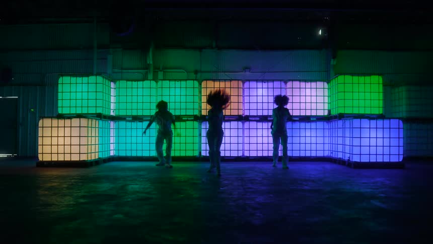 Three dancers perform a choreographed dance in front of illuminated cubes