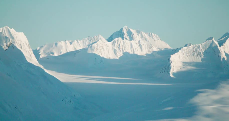 The video shows a wide static shot of a valley covered in snow and ice surrounded by sharp rocky snowcapped mountains the lighting is bright suggesting a sunny day in an arctic or highaltitude environment