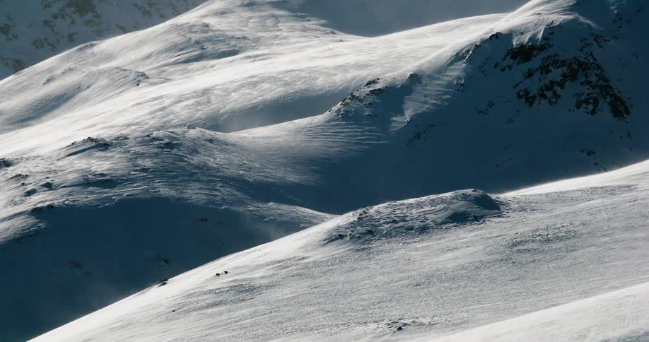 Windsculpted snow ripples across rolling alpine ridges with scattered rock outcrops casting long blue shadows under the low winter sun