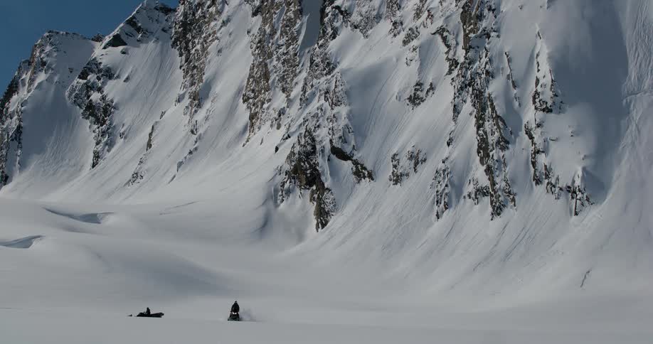 The video clip is an extreme long shot of a vast snowy and mountainous landscape at the base of a huge jagged mountain face two figures are dwarfed by the environment suggesting they are skiers or snowboarders in the backcountry