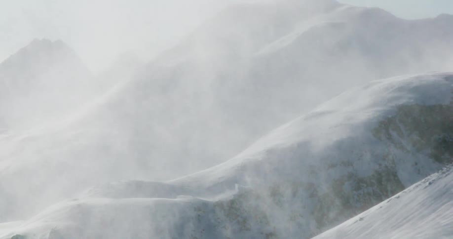 Wind whips plumes of snow across soft white mountain ridges veiling the distant peaks in a bright mist