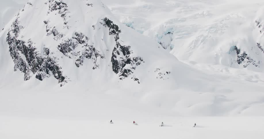 People cross a vast snowy landscape at the base of icy mountains