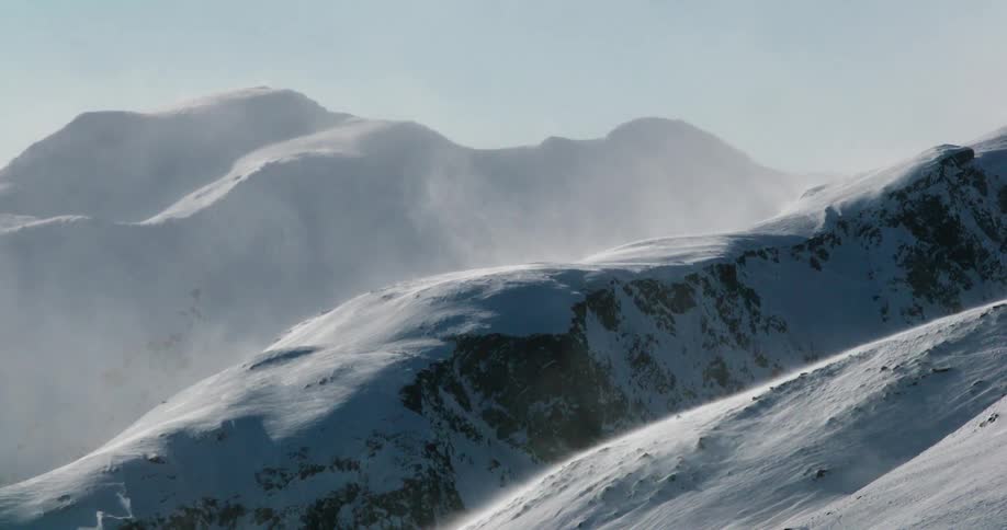 Wind lifts snow over a sunlit mountain range