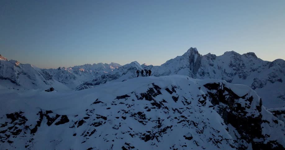 The video is an extreme long shot of a group of people standing on a snowy mountain ridge at sunset the figures are silhouetted against the bright sky and some appear to be holding skis or snowboards